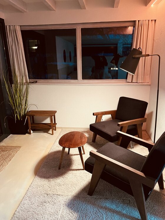 Sitzbereich im Pool House Zwei dunkle Sessel mit Holzgestell stehen vor einem Fenster neben einem kleinen runden Holztisch auf einem Teppich.