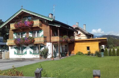 Alpines Wohnhaus mit Holzfassade, Balkonblumen und grün-weißen Fensterläden vor einer Wiese.