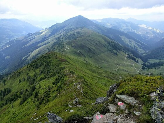 Blick von einem felsigen Berggipfel auf einen schmalen Wanderpfad, der über einen grünen Gebirgskamm führt.