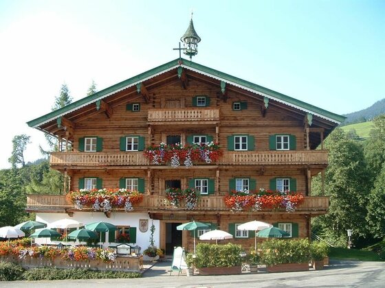 Gasthof Hallerwirt im Sommer Mehrstöckiges Holzhaus im Alpenstil mit Blumenschmuck an den Balkonen und einer Außenterrasse mit Sonnenschirmen.