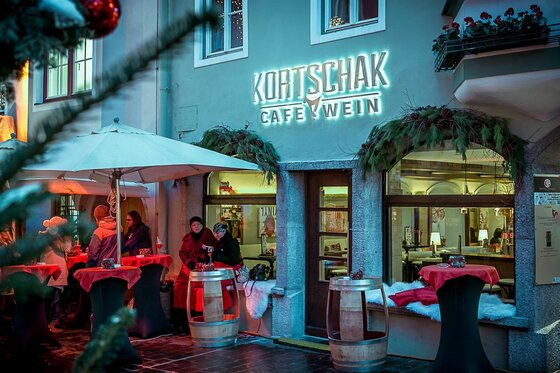 Kortschak Café & Wein Winter Außenansicht des Cafés Kortschak im Winter mit beleuchtetem Schriftzug, Stehtischen und Gästen.