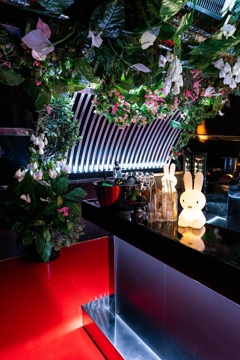 Moderne Bar mit dunklem Tresen, künstlicher Deckenbegrünung und leuchtenden Hasenfiguren.