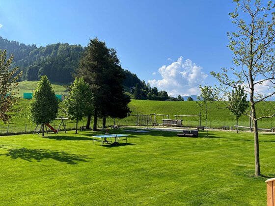 Schwimmbad Itter3 (c)ClarissaGünnewig Ein schöner, grüner Garten mit Bäumen und Spielgeräten. Im Hintergrund sind sanfte Hügel und ein blauer Himmel zu sehen.