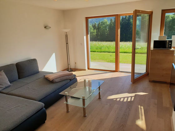 Ein einladendes Wohnzimmer mit einem grauen Sofa und einem Glastisch. Große Fenster bieten einen Blick auf die grüne Wiese draußen.