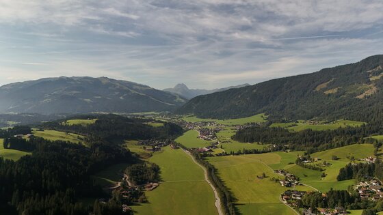 Bichlhof Coffee Ride Panorama Luftaufnahme eines Tals mit grünen Wiesen, kleinen Siedlungen und bewaldeten Bergen unter leicht bewölktem Himmel.