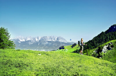 Wanderung am Berg Wanderin mit Rucksack auf einer grünen Bergwiese vor einem Gebirgspanorama mit Seilbahngondel.