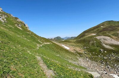 Schmaler Wanderpfad an einem grasbewachsenen Berghang unter blauem Himmel.
