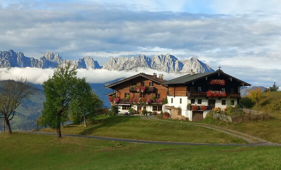 Herbst am Hinterkogel Zwei traditionelle Holzhäuser auf einer Wiese vor einem schroffen Gebirgsmassiv mit Wolkenband.