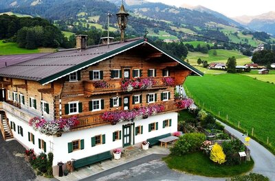 Ferienwohnung Kirchern Traditionelles Alpenhaus aus Holz und Stein mit Blumenschmuck an den Balkonen vor grünen Wiesen und Bergen.