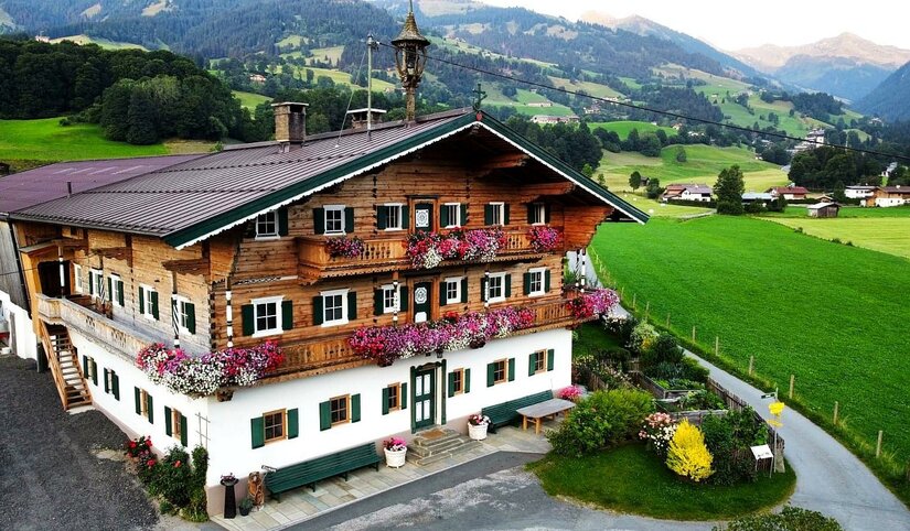 Ferienwohnung Kirchern Traditionelles Alpenhaus aus Holz und Stein mit Blumenschmuck an den Balkonen vor grünen Wiesen und Bergen.