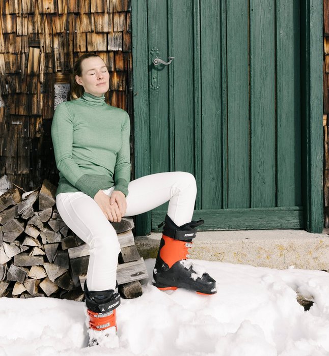 Eine Dame in Skibekleidung sitzt in der Sonne nach dem Skifahren an eine Holzhütte angelehnt