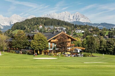 Rasmushof im Sommer Ein Holzgebäude am Rand eines Golfplatzes vor bewaldeten Hügeln und schroffen Berggipfeln.