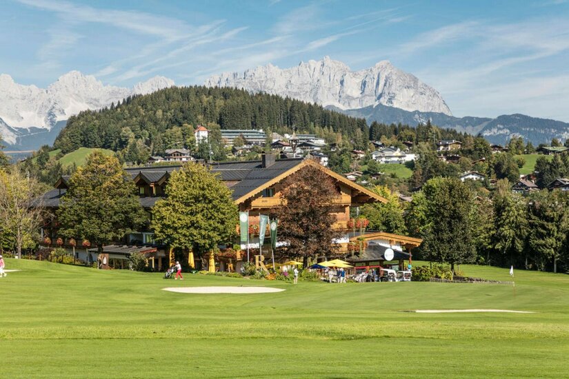 Rasmushof im Sommer Ein Holzgebäude am Rand eines Golfplatzes vor bewaldeten Hügeln und schroffen Berggipfeln.