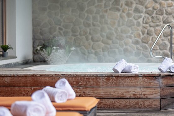 Wellnessbereich im Dorfresort Kitzbühel Dampfender Whirlpool mit Holzverkleidung, weißen Handtüchern und einer Natursteinwand im Hintergrund.