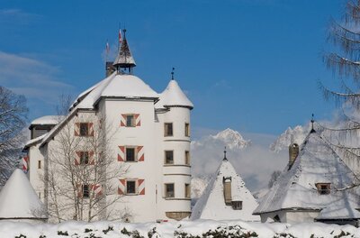 Weißes Schloss mit schneebedeckten Dächern und rot-weiß gestreiften Fensterläden vor Gebirgskulisse.