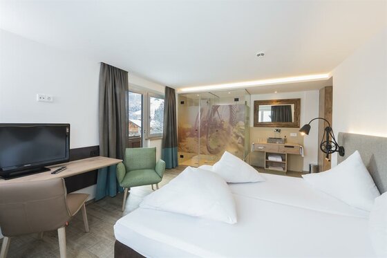 Modernes Doppelzimmer mit weißem Bett, Schreibtisch, Fernseher und offenem Badezimmerbereich.