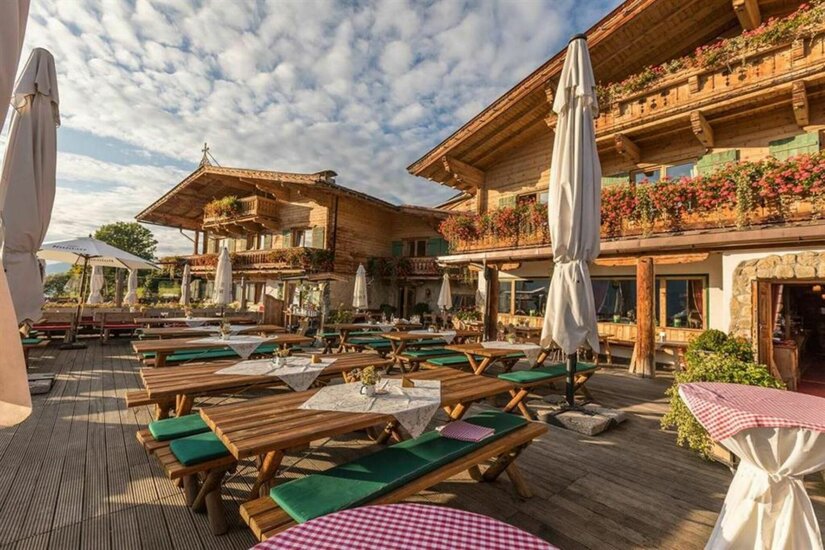 Sonnbergstuben Sommer Außenterrasse eines alpinen Holzgebäudes mit Holztischen, grünen Sitzkissen und Sonnenschirmen unter blauem Himmel.