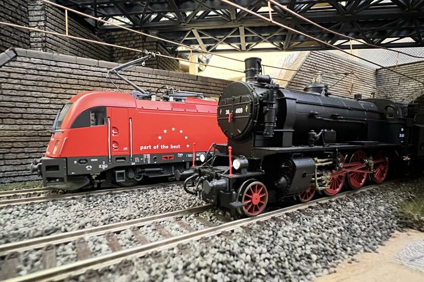Modelle einer roten Elektrolokomotive und einer schwarzen Dampflokomotive auf Schienen unter einer Brücke.