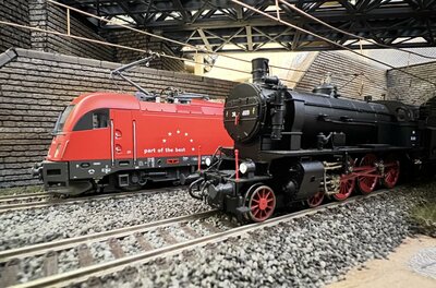 Modelle einer roten Elektrolokomotive und einer schwarzen Dampflokomotive auf Schienen unter einer Brücke.