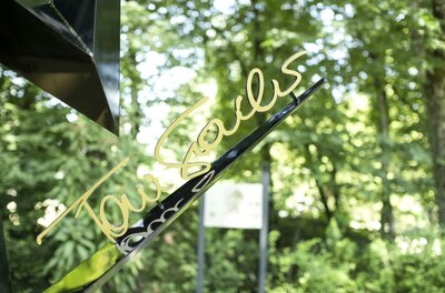 Ein elegantes Schild mit der Schrift "Tourists" in goldener Farbe, das vor einer grünen, bewaldeten Kulisse steht. Der ruhige Hintergrund sorgt für eine angenehme Atmosphäre.
