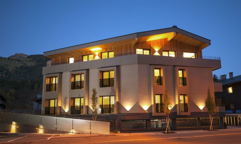 Beleuchtetes, modernes Apartmenthaus in der Dämmerung mit Holzfassade im Dachgeschoss.