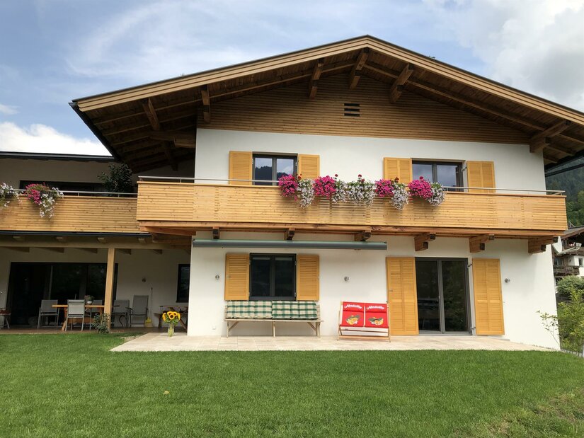 Zweistöckiges alpines Wohnhaus mit Holzfassade, Balkonblumen und einer Terrasse mit Sitzgelegenheiten.