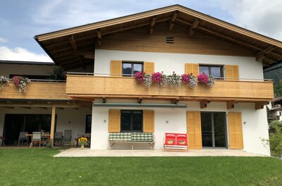 Haus Staffa Außenansicht Zweistöckiges alpines Wohnhaus mit Holzfassade, Balkonblumen und einer Terrasse mit Sitzgelegenheiten.