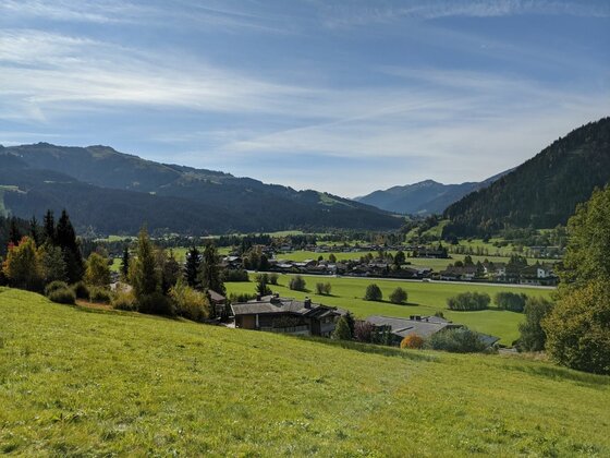 Blick auf das Alpental Blick über eine grüne Wiese auf ein Alpendorf im Tal vor bewaldeten Bergen unter blauem Himmel.