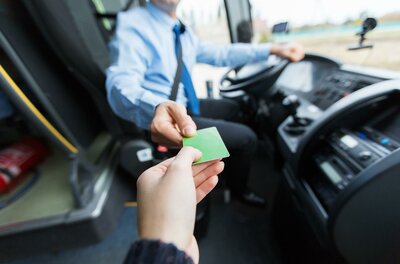 Ein Fahrgast gibt dem Busfahrer eine grüne Fahrkarte. Der Fahrer sitzt am Steuer des Busses.