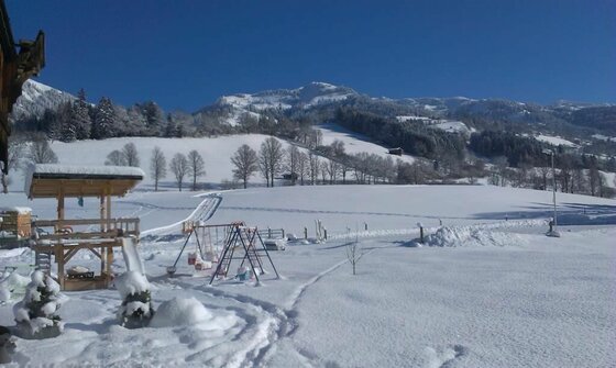 Fichterhof Kitzbühel Winterlandschaft Verschneiter Spielplatz vor einer alpinen Bergkulisse unter blauem Himmel.