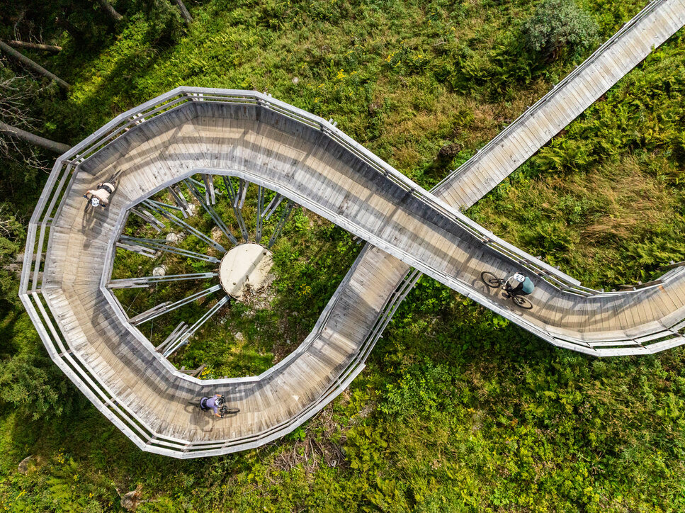 Eine geschwungene Holzbrücke am Sonnenrast Trail in Kitzbühel, umgeben von üppigem Grün, während Mountainbiker die Strecke nutzen.