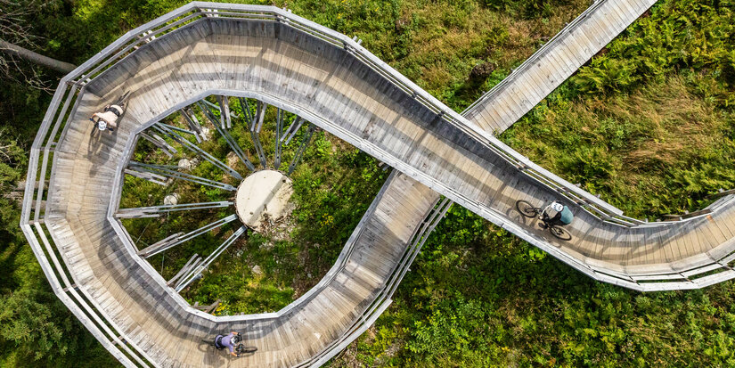 In Kitzbühel, two cyclists ride across a curved wooden bridge in a green, wooded setting.