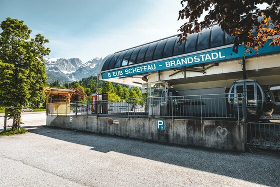 Eine Seilbahnstation in der Bergregion mit Blick auf die verschneiten Berge. Die Umgebung ist grün und einladend, ideal für Ausflüge in der Natur.