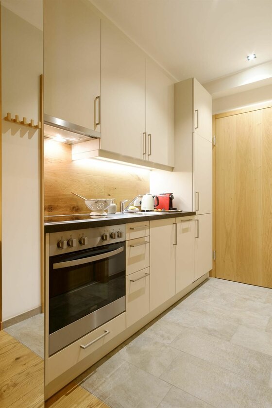 Küchenzeile Apartment Helene Moderne Küchenzeile in Beige mit Holzrückwand, Herd und hellen Bodenfliesen.