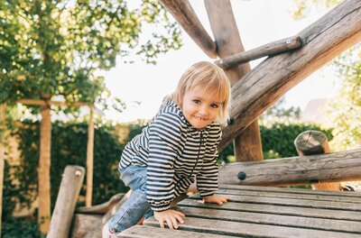 Kind auf Holzspielplatz Ein Kleinkind in gestreifter Jacke klettert auf einem Holzspielgerät im Freien.