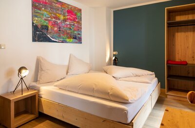Modernes Doppelbett aus Holz mit weißer Bettwäsche in einem hellen Zimmer mit buntem Wandbild.