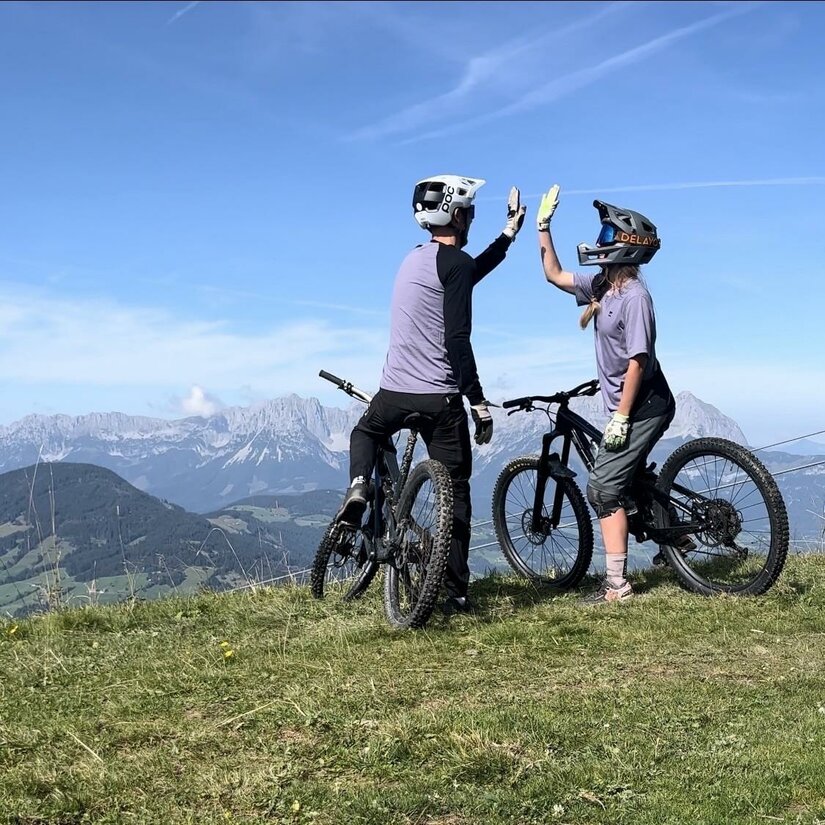 Mountainbiker am Gipfel Zwei Mountainbiker geben sich auf einem Berggipfel vor einem Alpenpanorama ein High-Five.