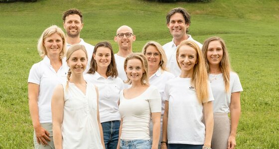 Team der Rehabilitation Aurach Zehn Personen in weißer Kleidung stehen lächelnd vor einer grünen Wiese im Freien.