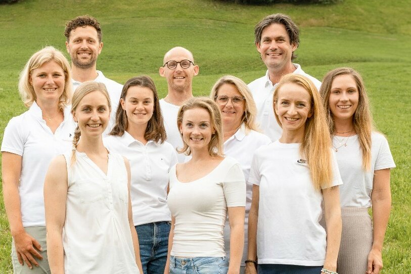 Team der Rehabilitation Aurach Zehn Personen in weißer Kleidung stehen lächelnd vor einer grünen Wiese im Freien.