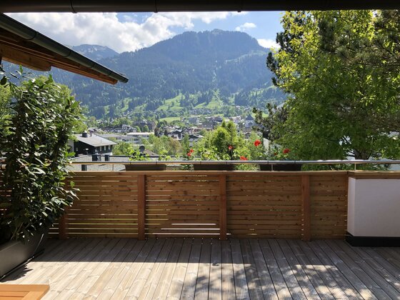 Blick von einer Holzterrasse auf ein Bergdorf und bewaldete Hänge unter blauem Himmel.