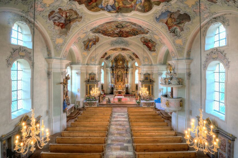 Eine wunderschöne Kircheninneneinrichtung mit gewölbter Decke und kunstvollen Fresken. Das Interieur ist hell erleuchtet und zeigt Holzbänke sowie goldene Kronleuchter.