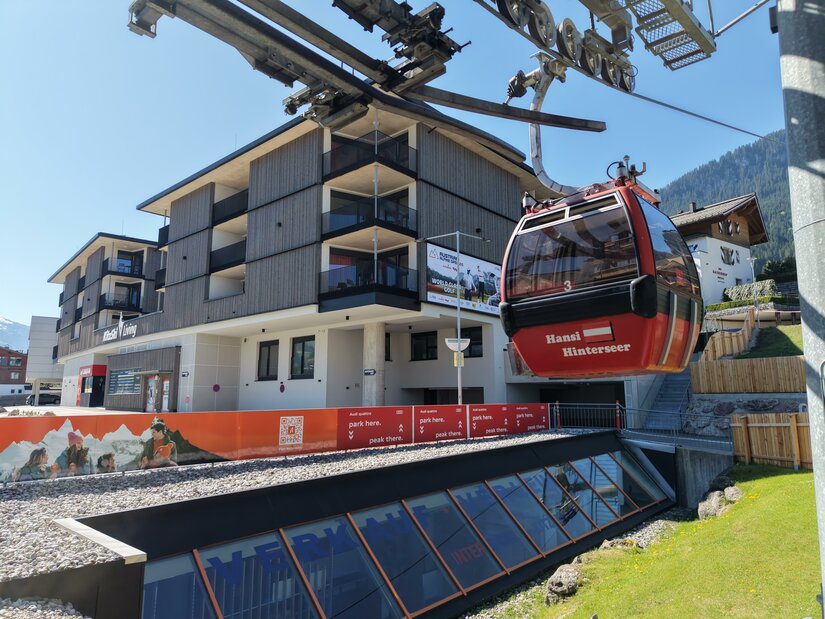 Eine moderne Gondelbahn transportiert Passagiere zu einem Gebirgshotel. Im Hintergrund sind die Berge und der blaue Himmel zu sehen.
