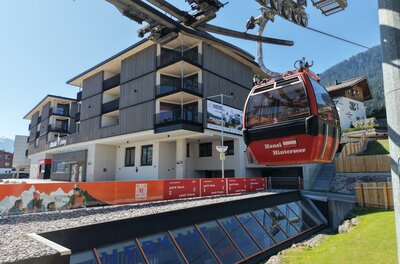DJI_20260424062103_0287_D Eine moderne Gondelbahn transportiert Passagiere zu einem Gebirgshotel. Im Hintergrund sind die Berge und der blaue Himmel zu sehen.