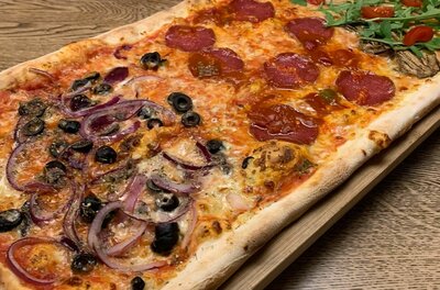 Pizza-Variation im Don Luigi Längliche Pizza mit verschiedenen Belägen wie Salami, Oliven, roten Zwiebeln und Rucola auf einem Holzbrett.