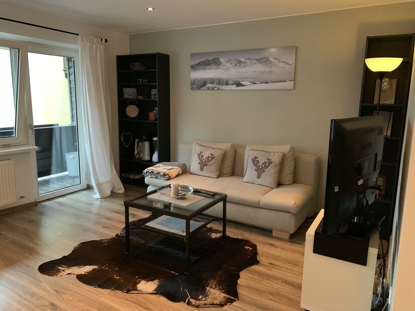 Wohnzimmer mit hellem Sofa, Glastisch, Kuhfellteppich und einem Bergbild an der Wand.