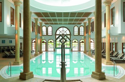 Hallenbad mit Säulen, Liegestühlen und einer Metallskulptur im Vordergrund.