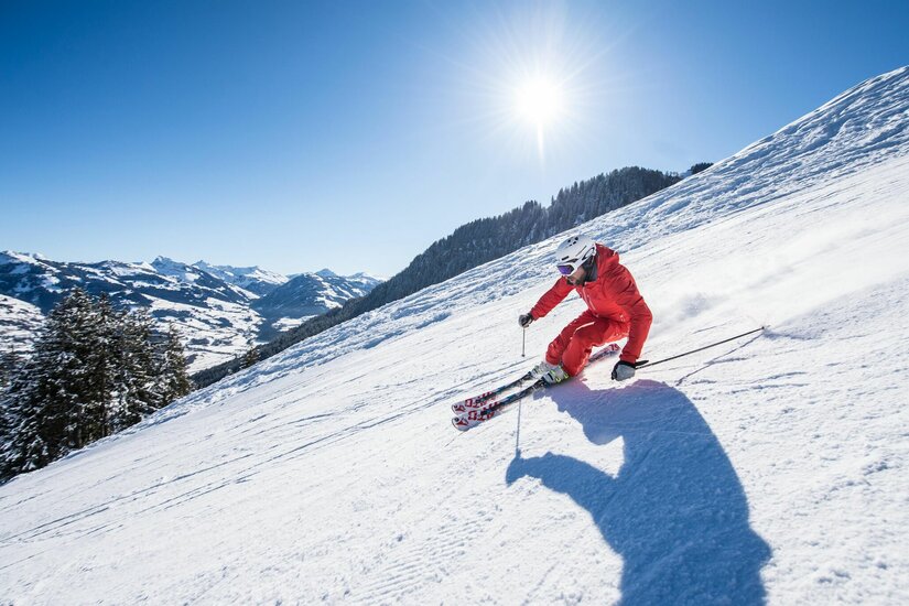 Snowsports Kitzbühel Skifahrer in roter Kleidung fährt eine präparierte Piste bei Sonnenschein hinunter.
