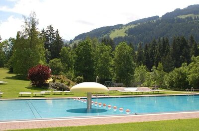 Freibad mit blauem Wasserbecken, Wasserpilz und Liegewiese vor bewaldeten Bergen.