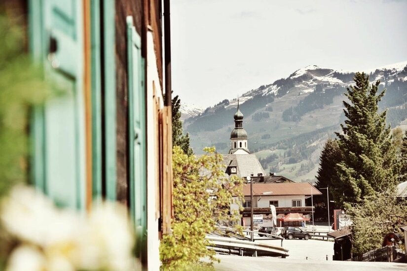 Blick auf eine Dorfkirche mit Zwiebelturm vor schneebedeckten Bergen, eingerahmt von einer Hausfassade.