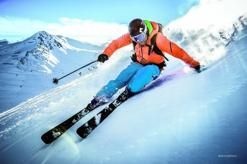 Skifahrer bei Abfahrt Skifahrer in orangefarbener Jacke und blauer Hose fährt eine steile, verschneite Piste hinunter.